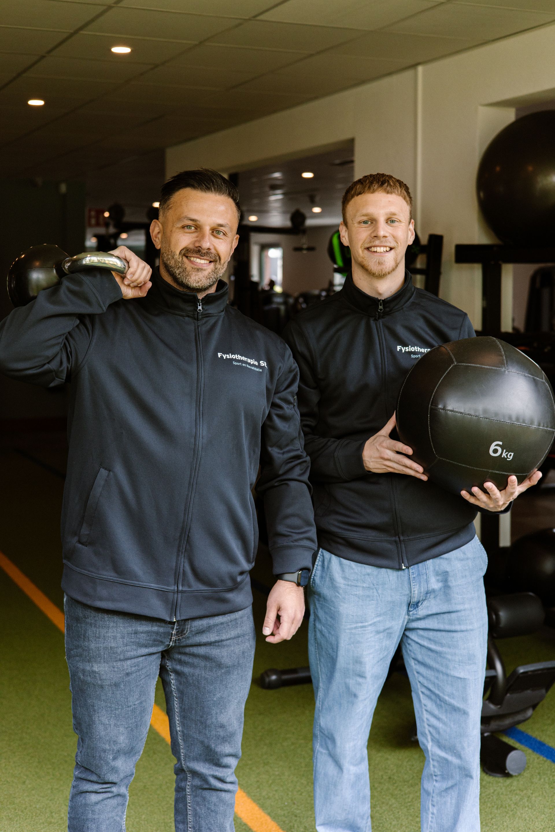 Twee mannen in zwarte jassen poseren in een sportschool, een van hen houdt een medicijnbal vast.