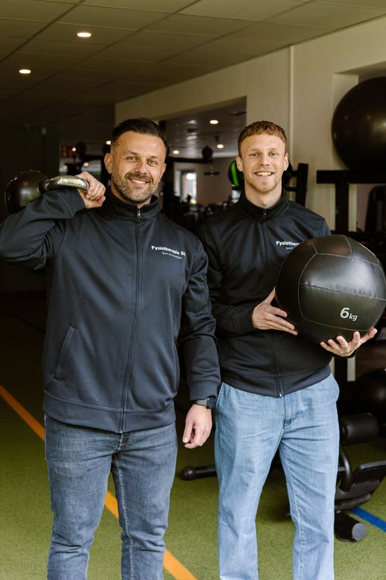 Twee mannen in zwarte jassen poseren in een sportschool, een van hen houdt een medicijnbal vast.