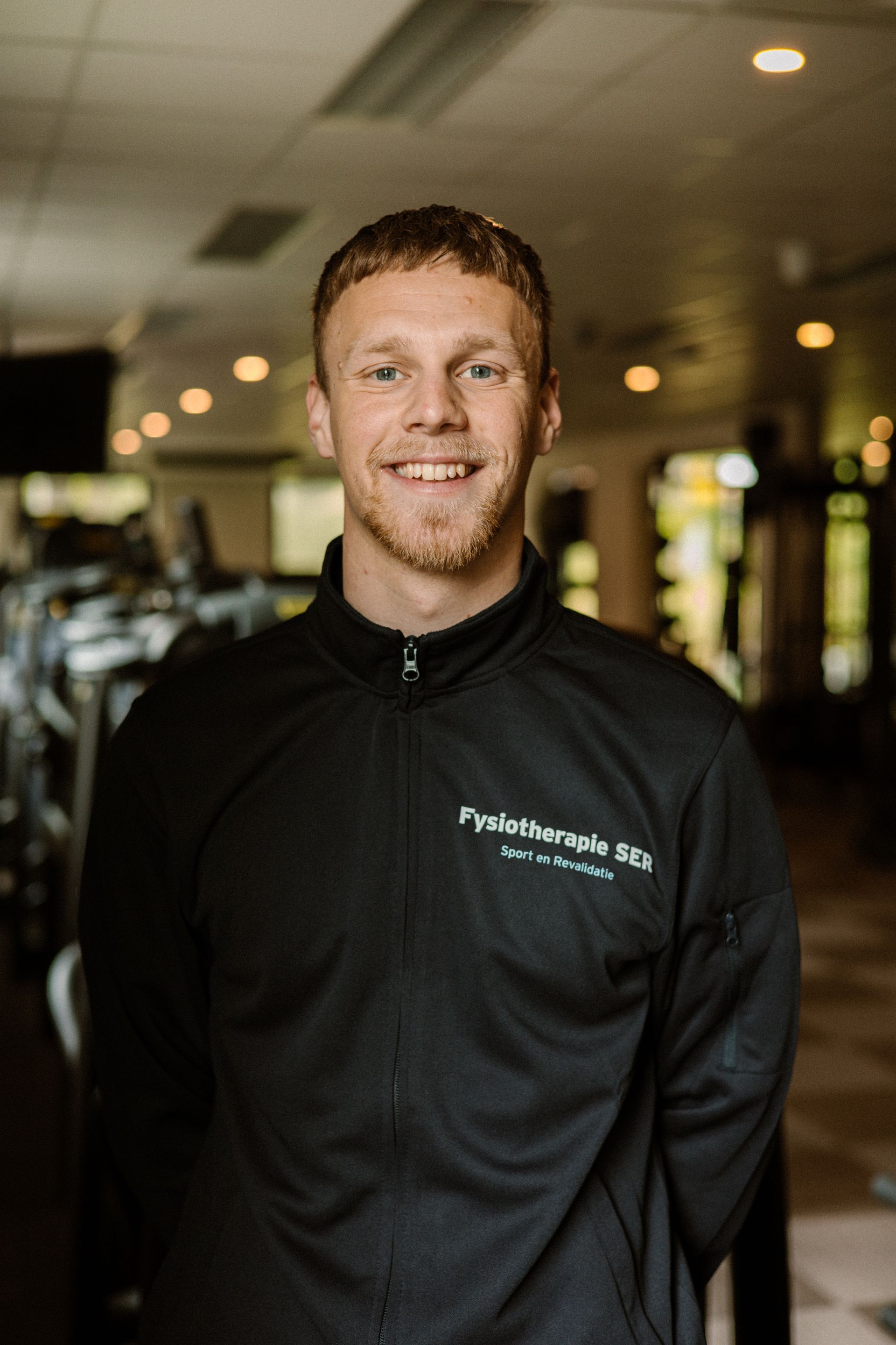 Een lachende man in een zwart vest met rits staat in de gang van een sportschool.