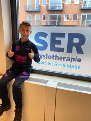 Persoon steekt twee duimen omhoog naast een SER-fysiotherapiebord in een raam.