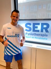 Een man staat binnen met een trui in zijn hand voor een bord van SER Fysiotherapie.