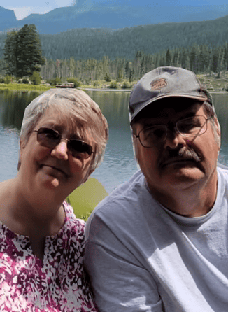 Two people posing by a lake with forested mountains in the background.