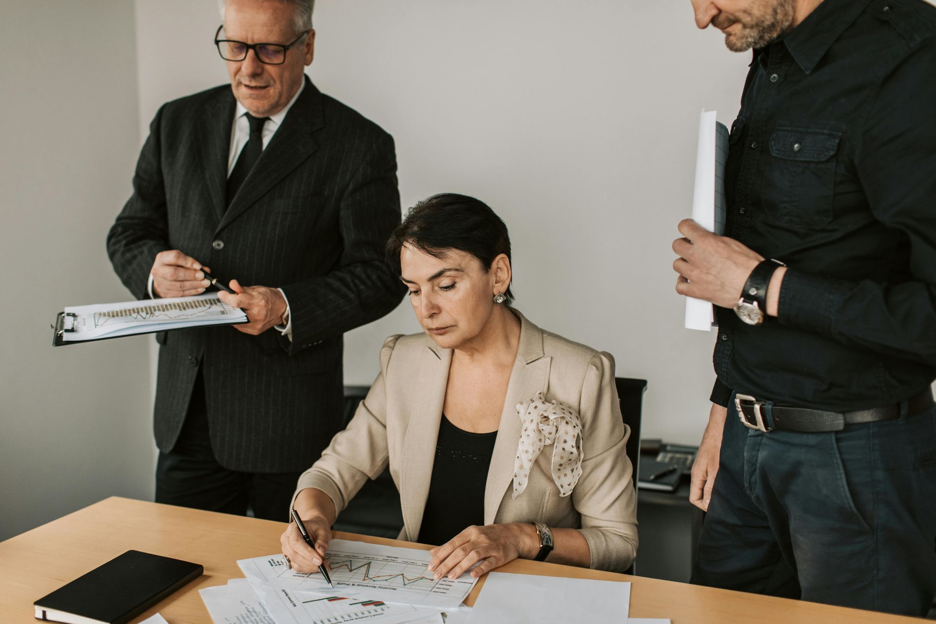 Three coworkers review documents at a desk in a meeting room, with one seated and two standing nearby.