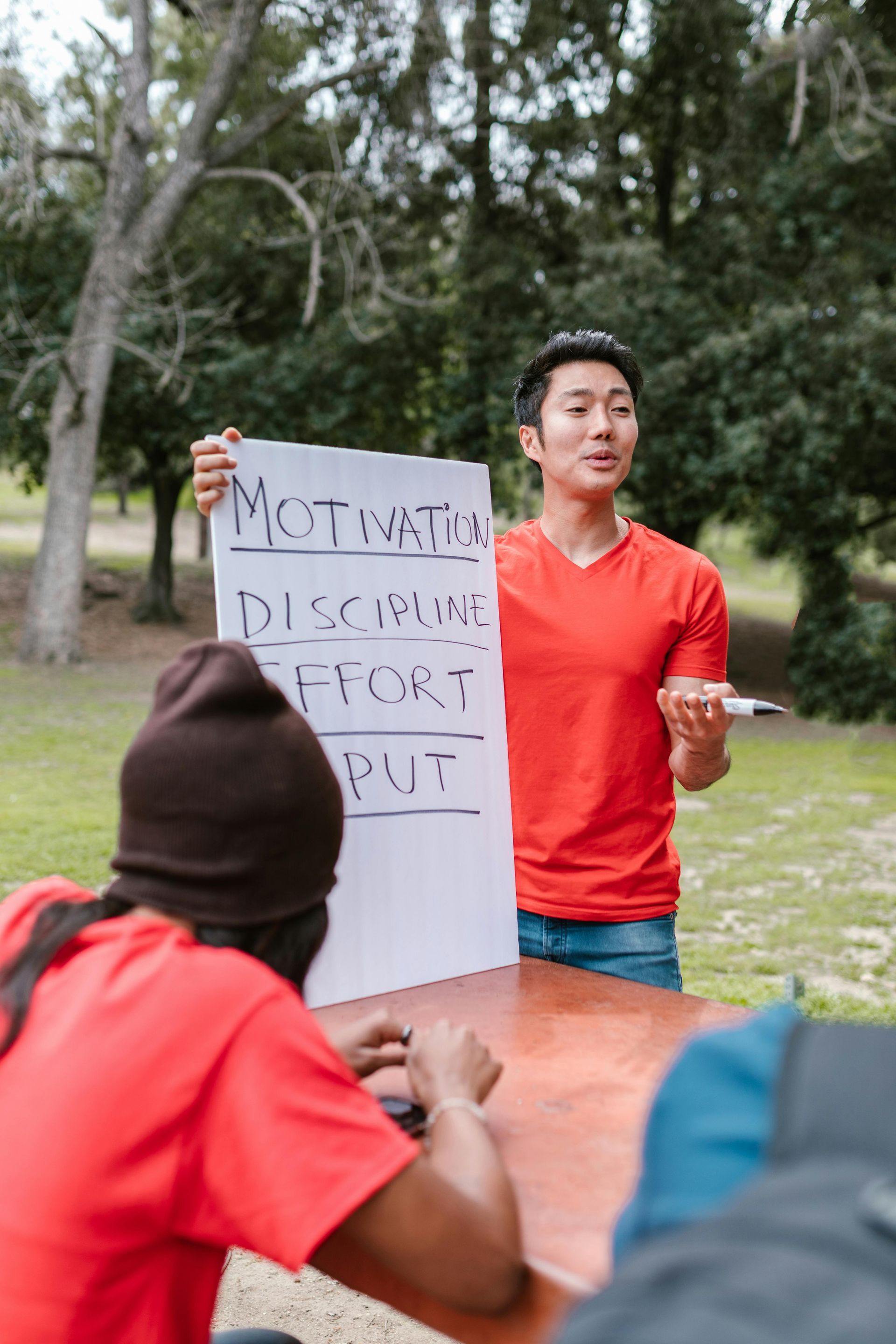 Person in red shirt holding a handwritten sign during an outdoor discussion, with others seated nearby