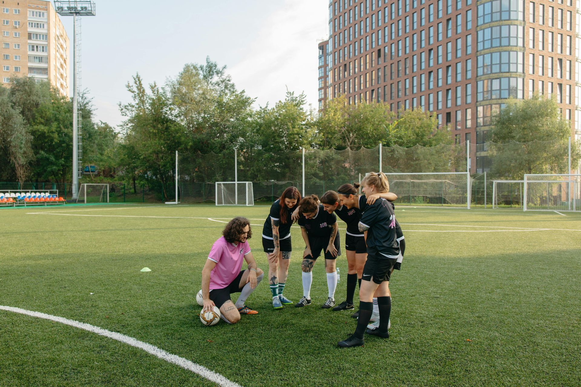 Youth soccer players huddle on a field while a coach kneels beside them near the sideline.
