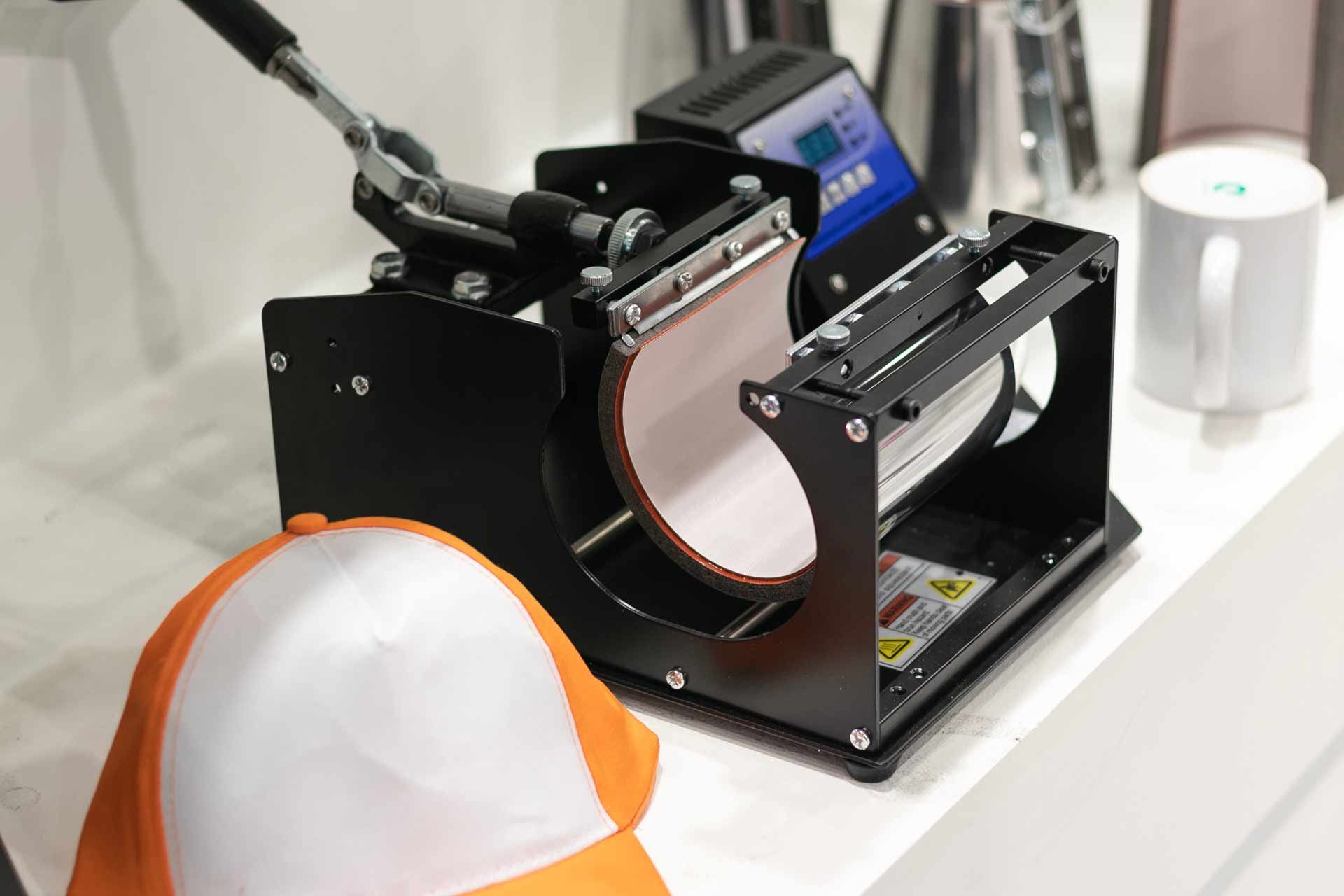 Sublimation heat press machine with orange-and-white cap being pressed in a workshop setting