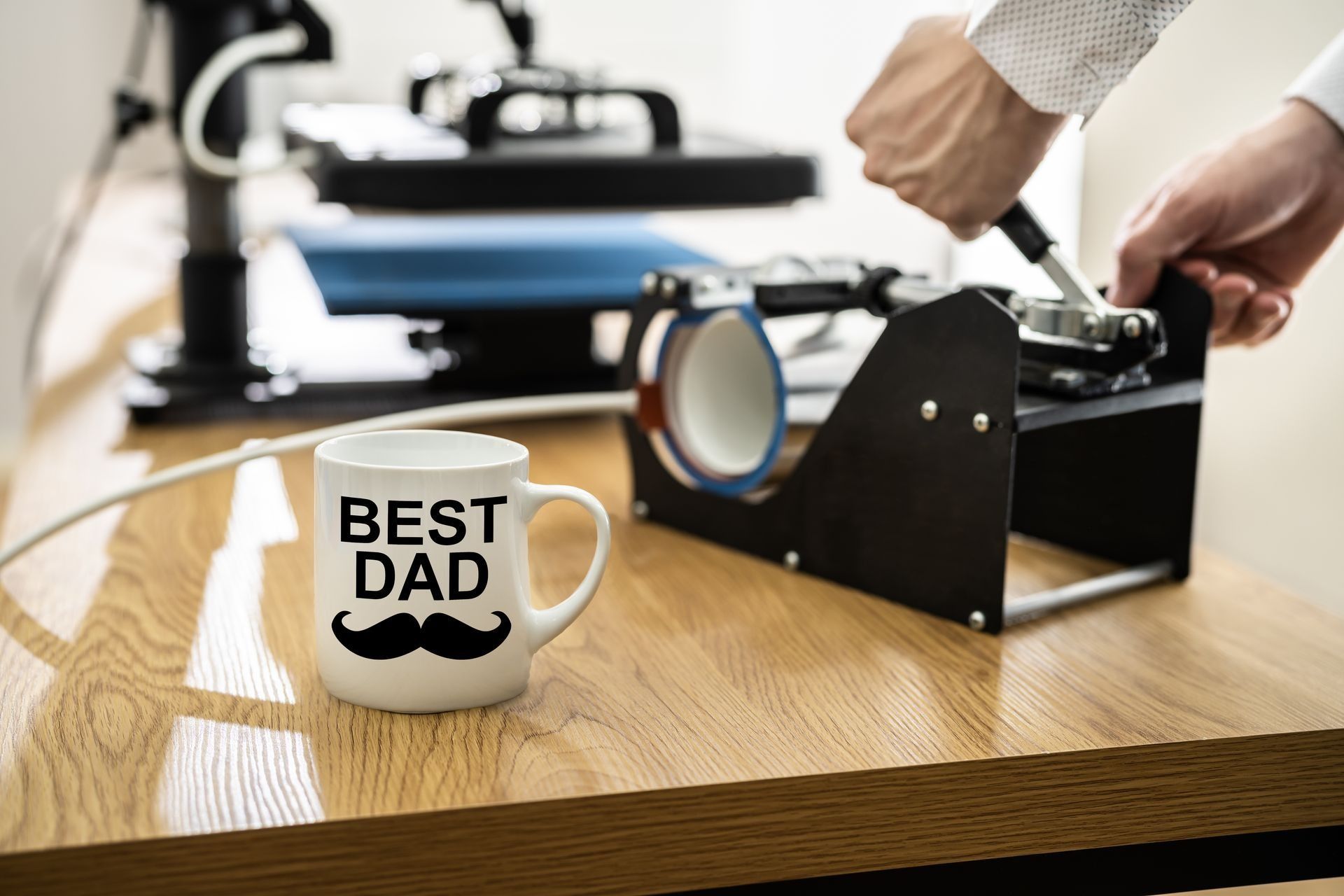 “Best Dad” mug beside a black heat press on a wooden table, with hands adjusting the machine