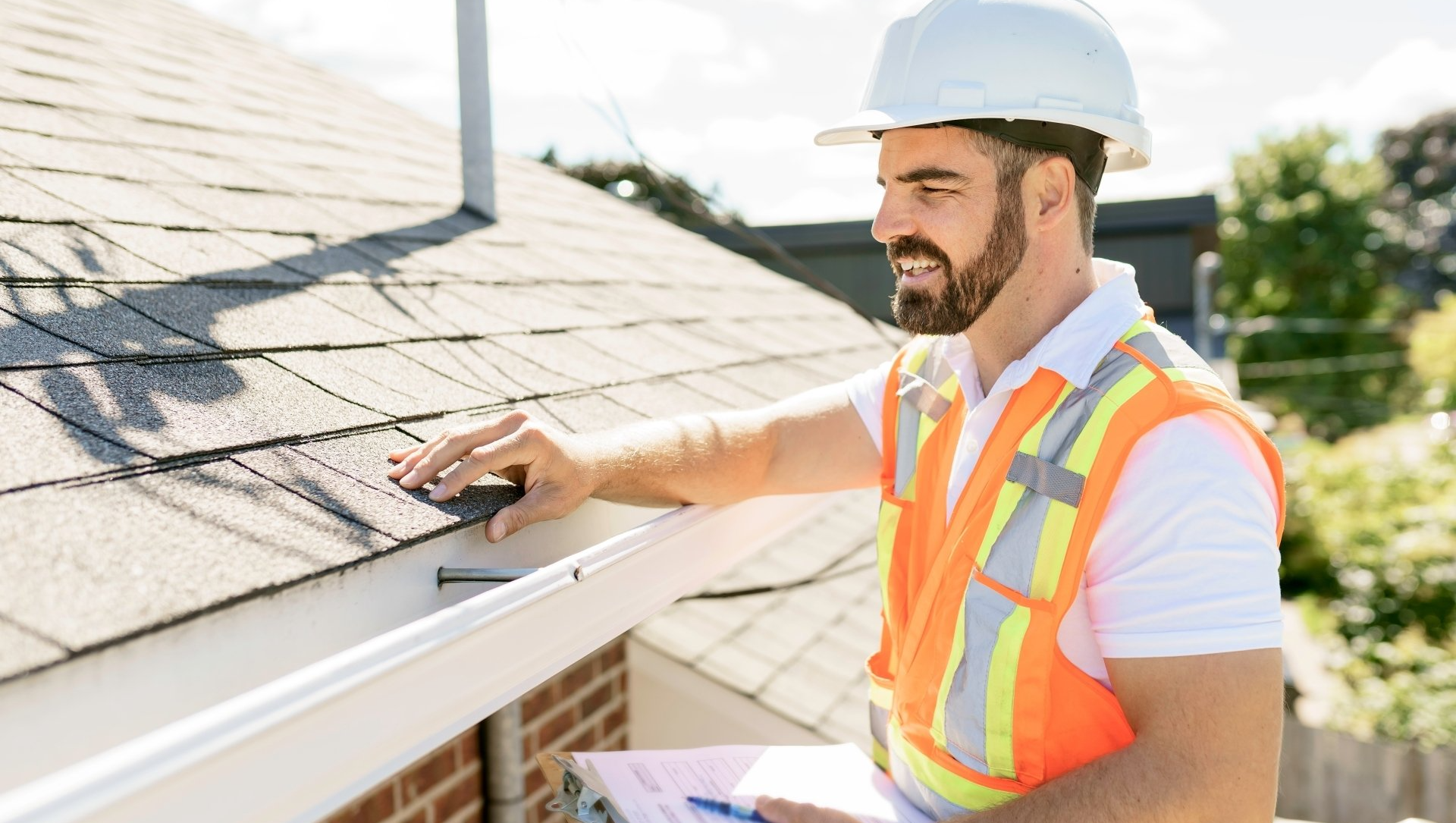 A man in a hard hat and orange vest is standing on a roof looking at a gutter.
