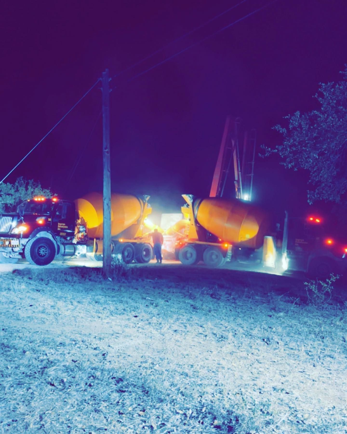 Two concrete mixer trucks at night, under lights. Workers are present, near equipment.