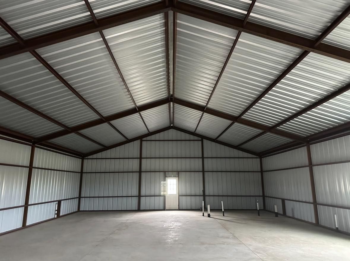 Interior view of an empty metal-framed building with a high, peaked ceiling. Brown framing, white walls, and a concrete floor.