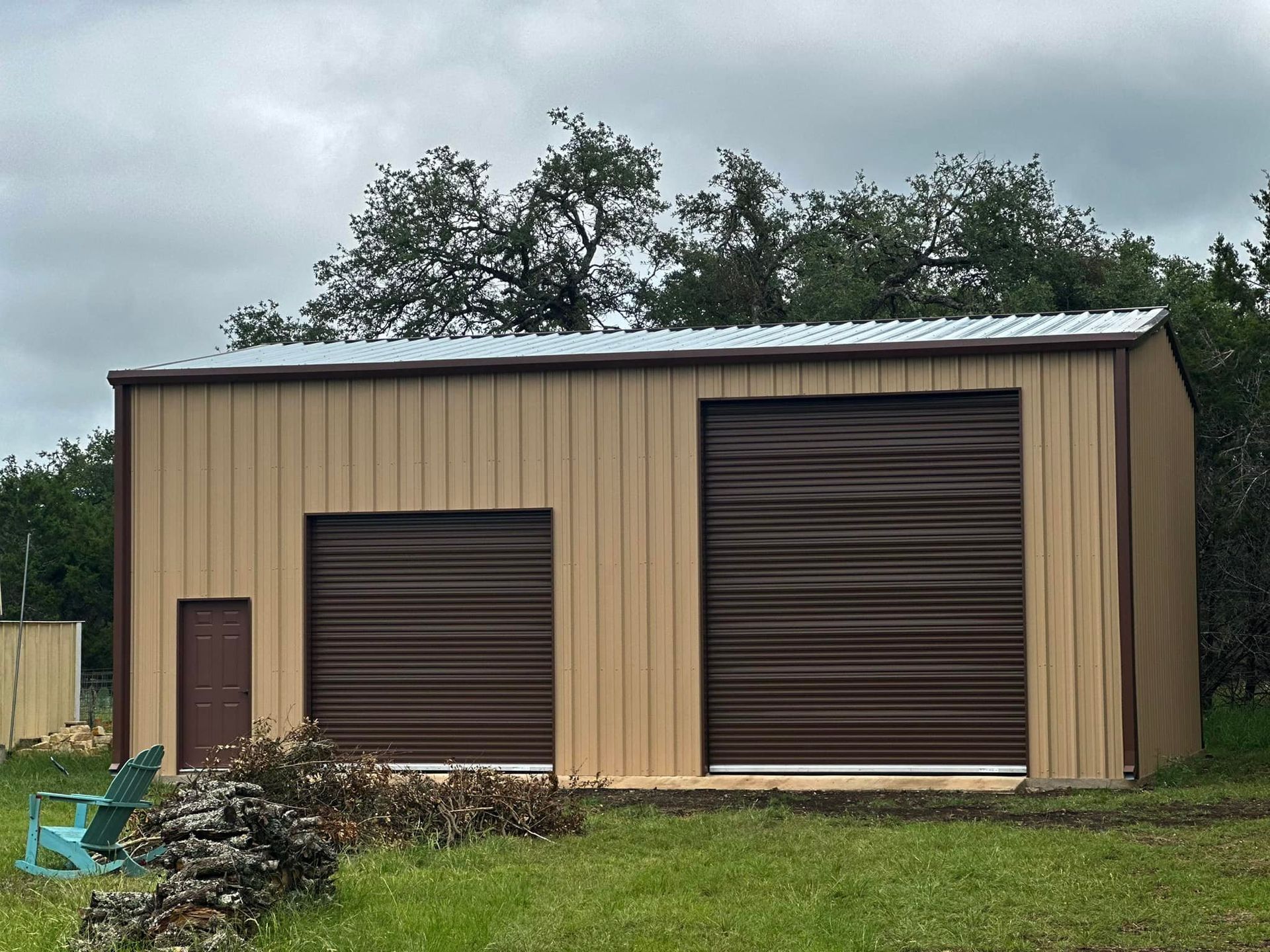 Tan metal building with brown trim and roll-up doors, on green grass under a cloudy sky.
