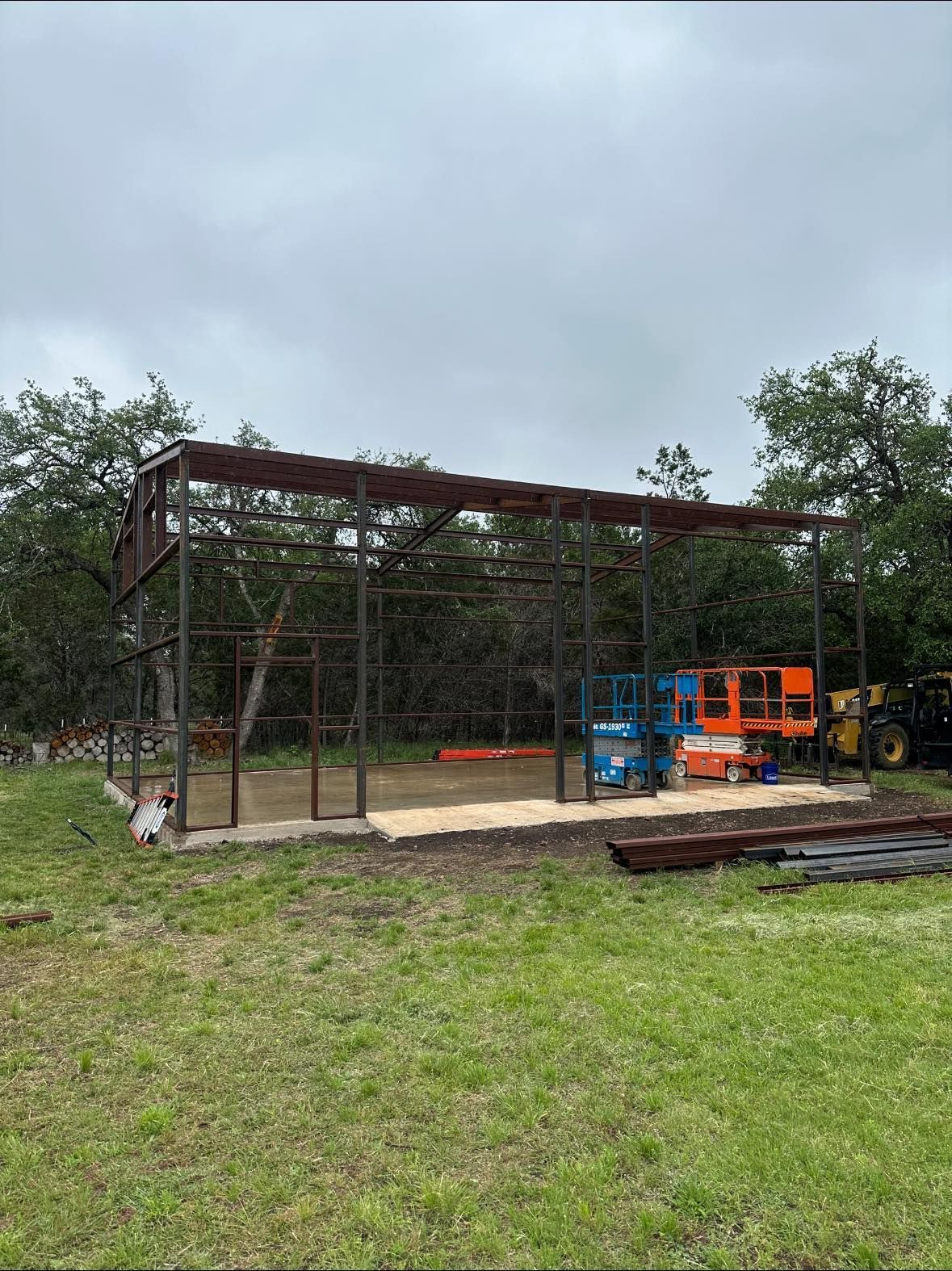 Steel-framed building under construction on a grassy field; lifts and tractor visible; trees in background.