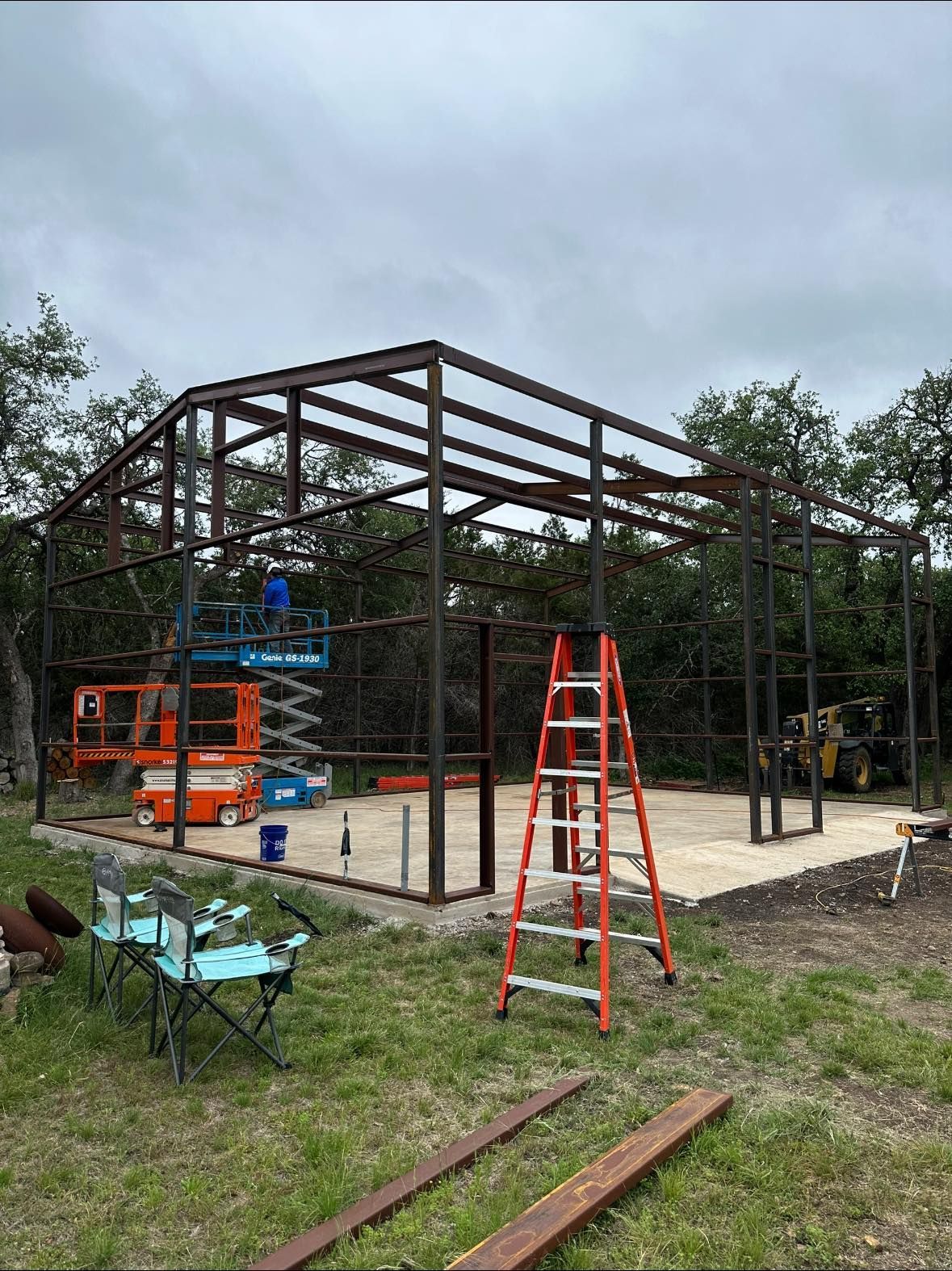 Steel frame construction of a building on a concrete foundation; a scissor lift and ladder are present.