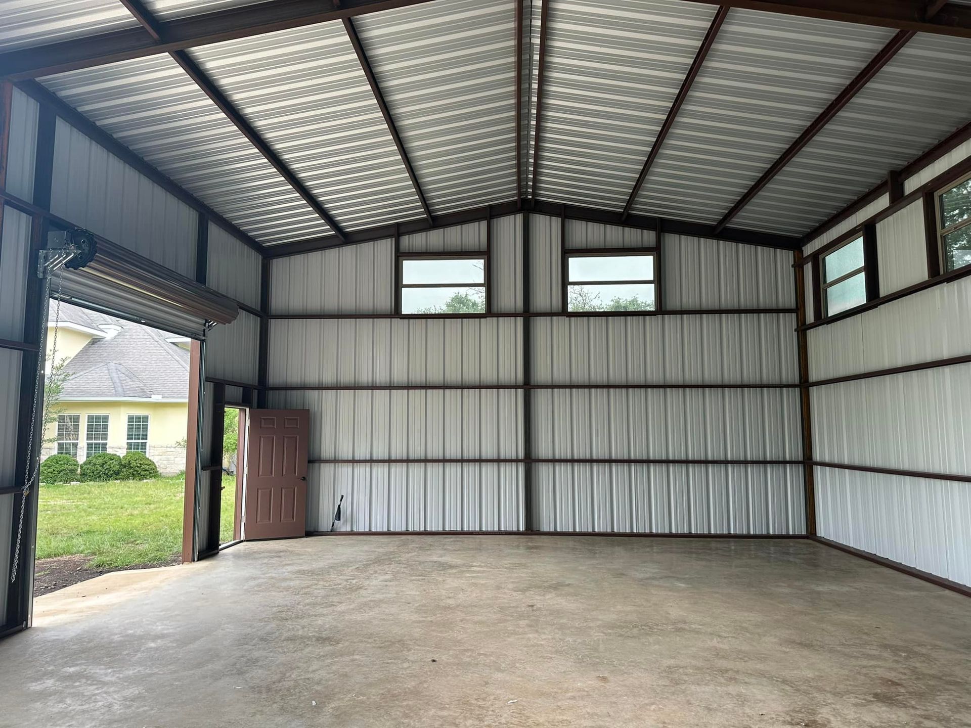 Inside of an empty metal shed with a concrete floor, two windows, and an open garage door.