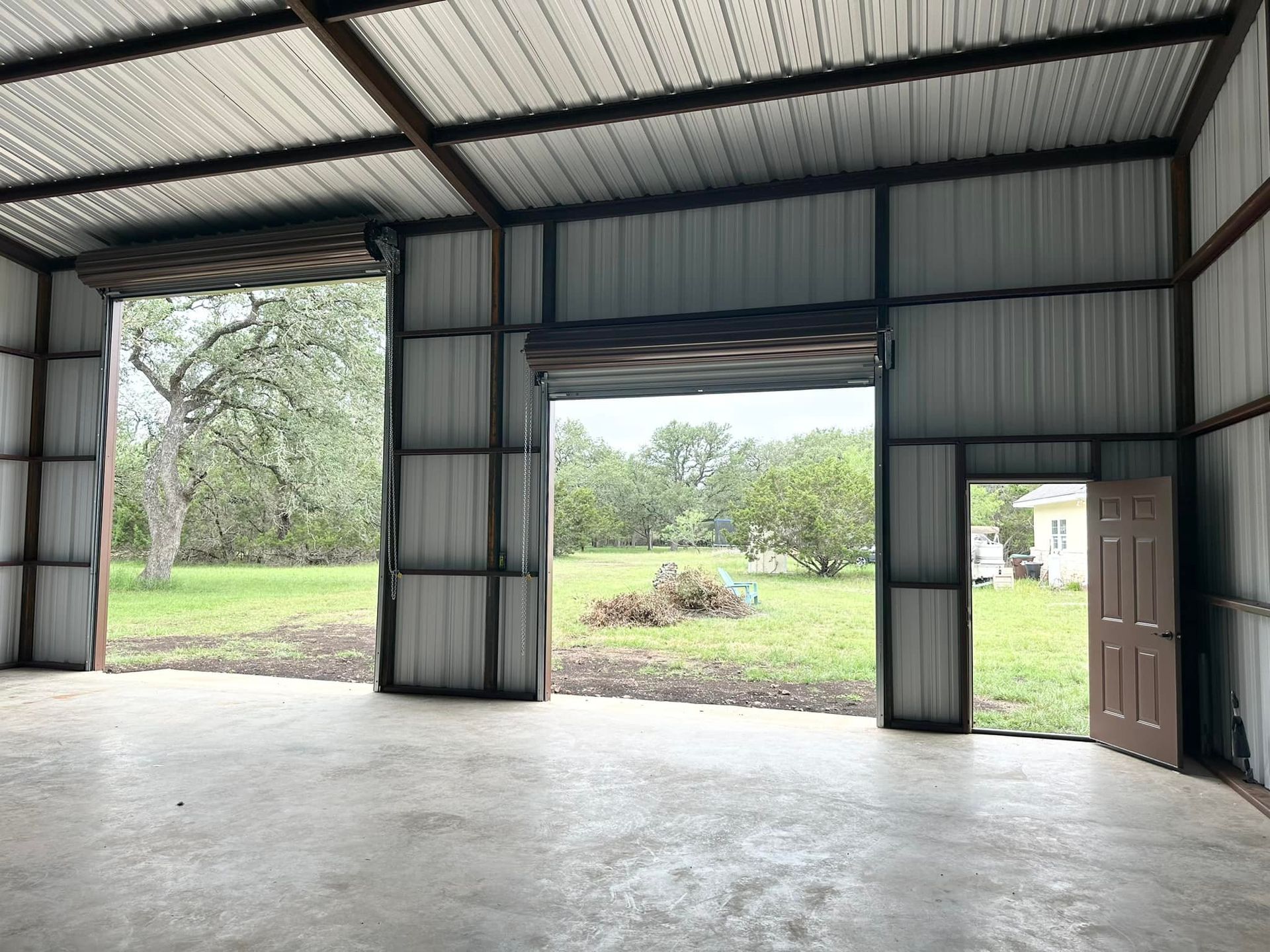 Interior of a metal building with open roll-up doors facing a grassy field and trees.