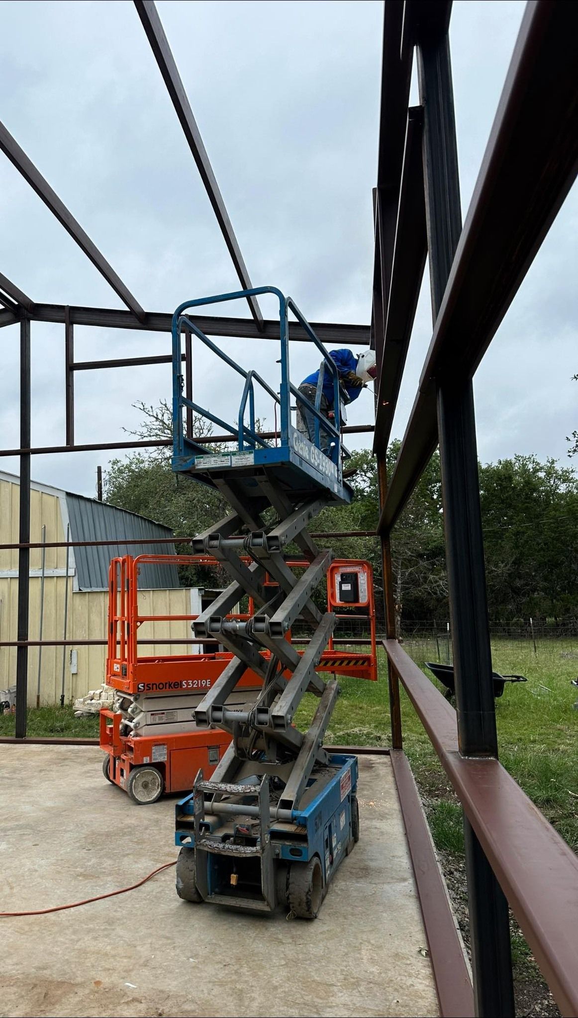 Construction worker on lift welding metal framing outdoors. Orange and blue scissor lifts on concrete.