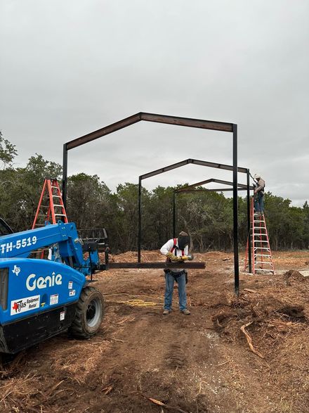 Construction of a steel-framed structure with a lift. Workers and a Genie lift on a muddy worksite with trees in the background.