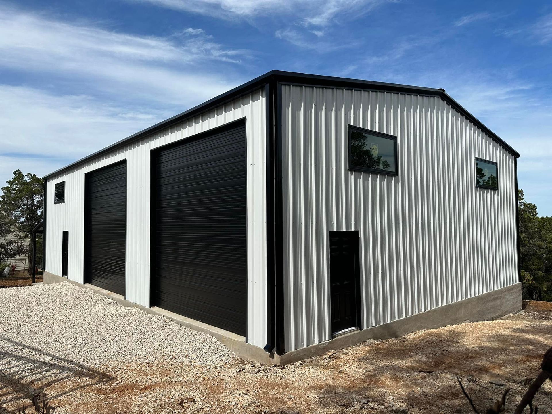 Metal building with two black garage doors, white siding, and two windows. Set on gravel.