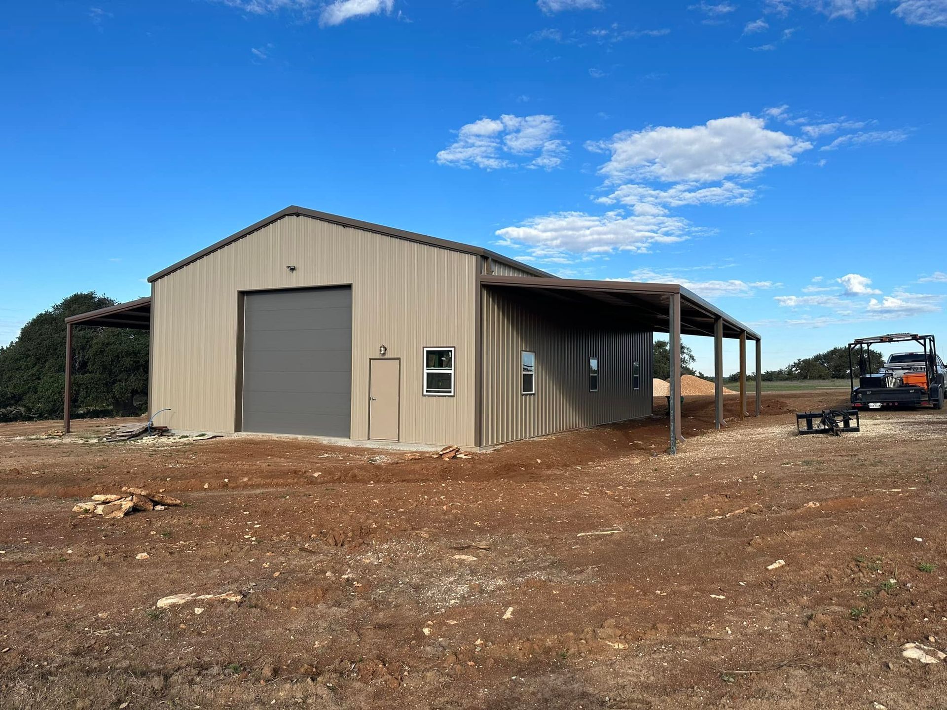 Tan metal building with attached covered patio, a garage door, and a small window on a brown dirt lot.