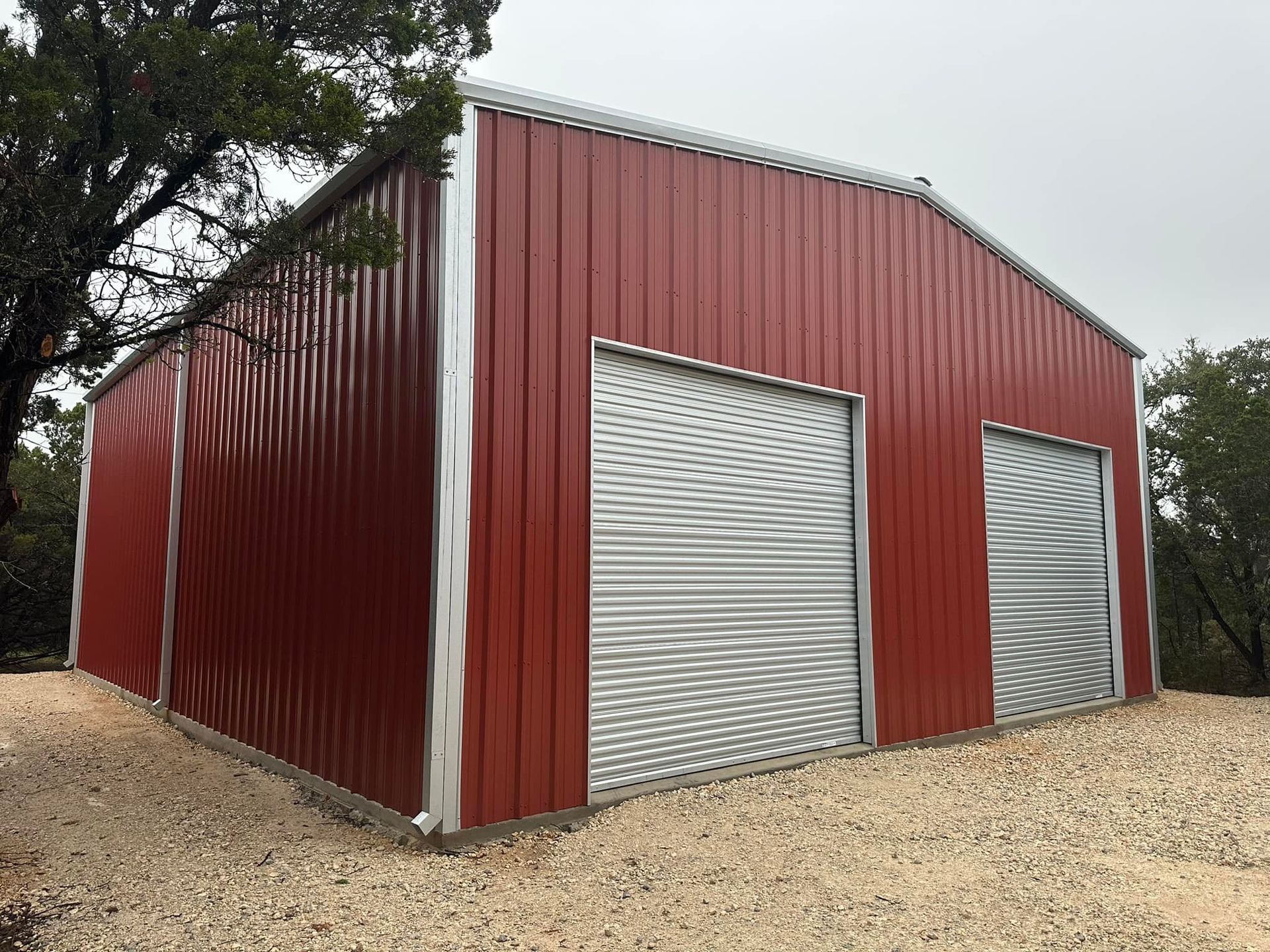 Red metal building with roll-up doors, on a gravel base, set against trees.