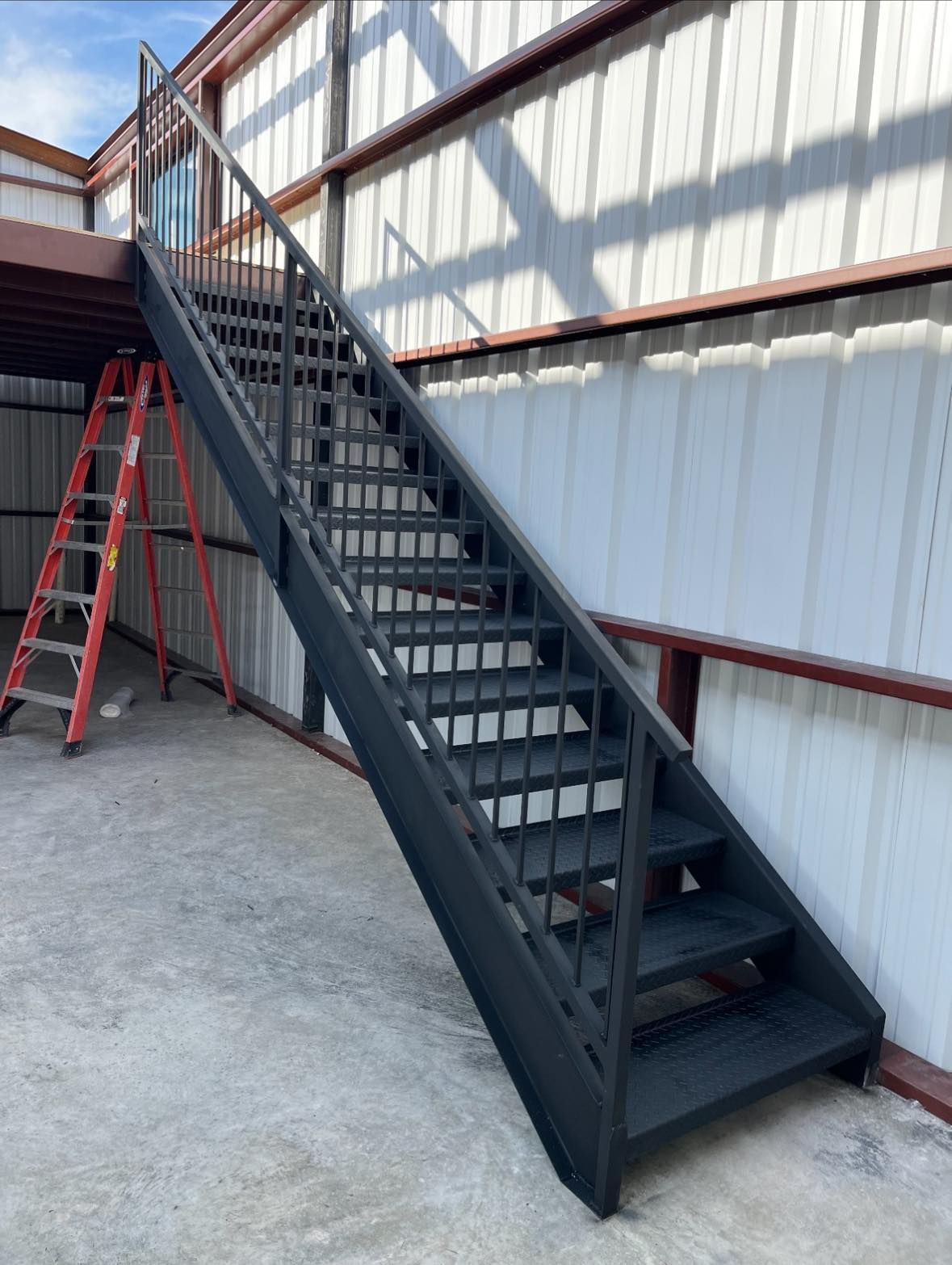 Black metal staircase leading to a mezzanine inside a warehouse; red ladder to the side.