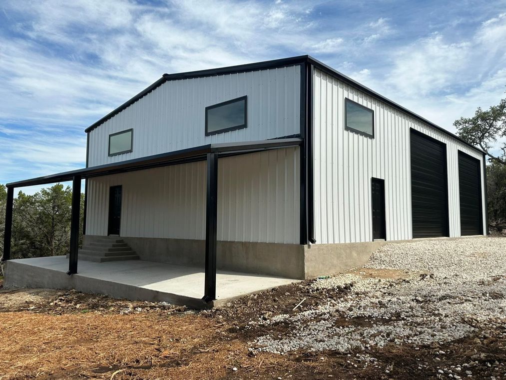 White metal building with black trim, porch, and rolling doors. Set on a gravel lot under a blue sky.