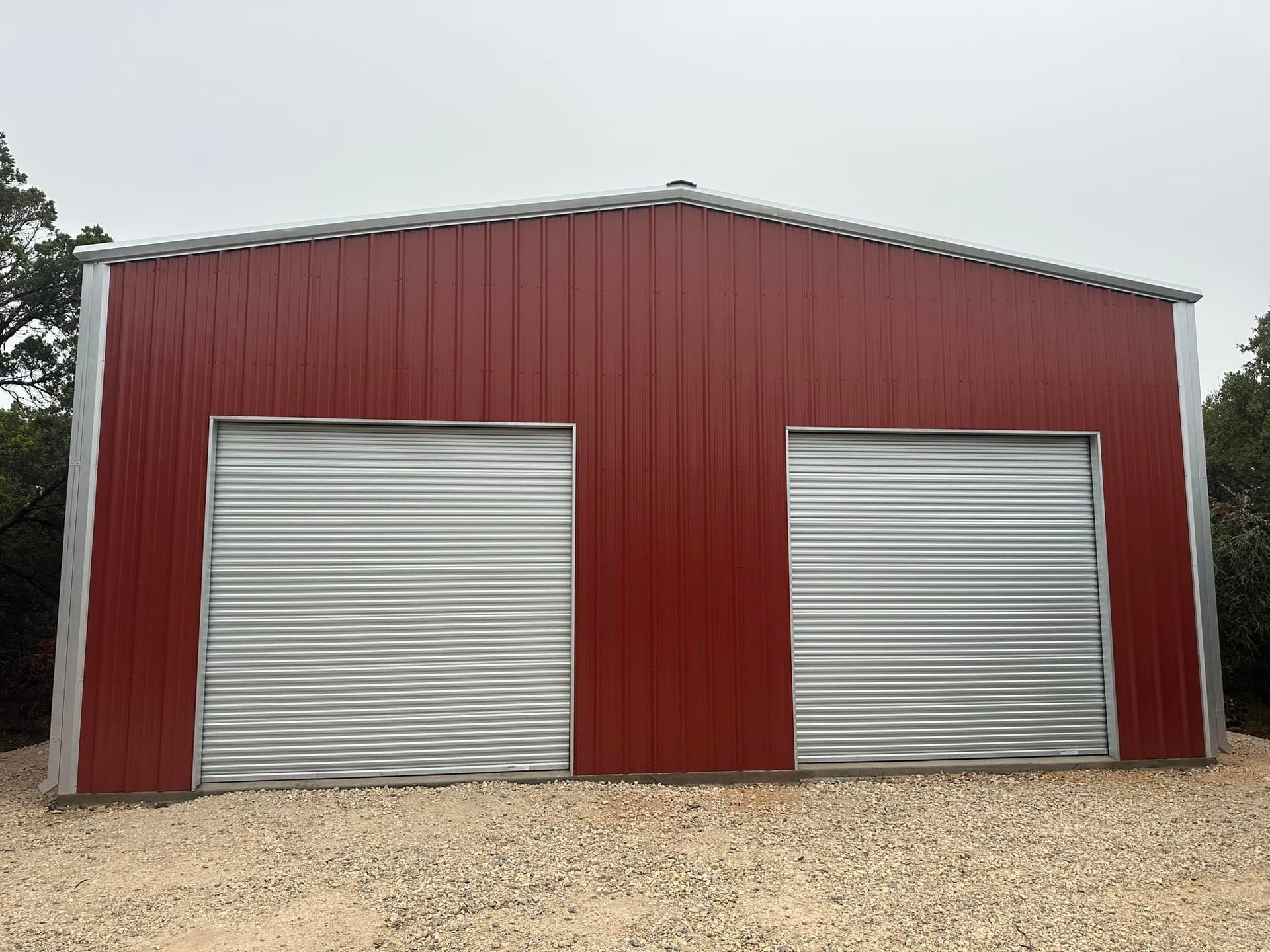 Red metal garage with two roll-up doors, set on gravel, under a gray sky.