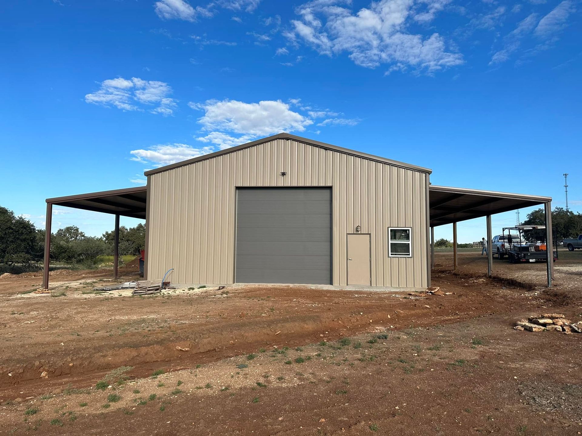 Tan metal building with attached carports, gray garage door, and a small window against a blue sky.