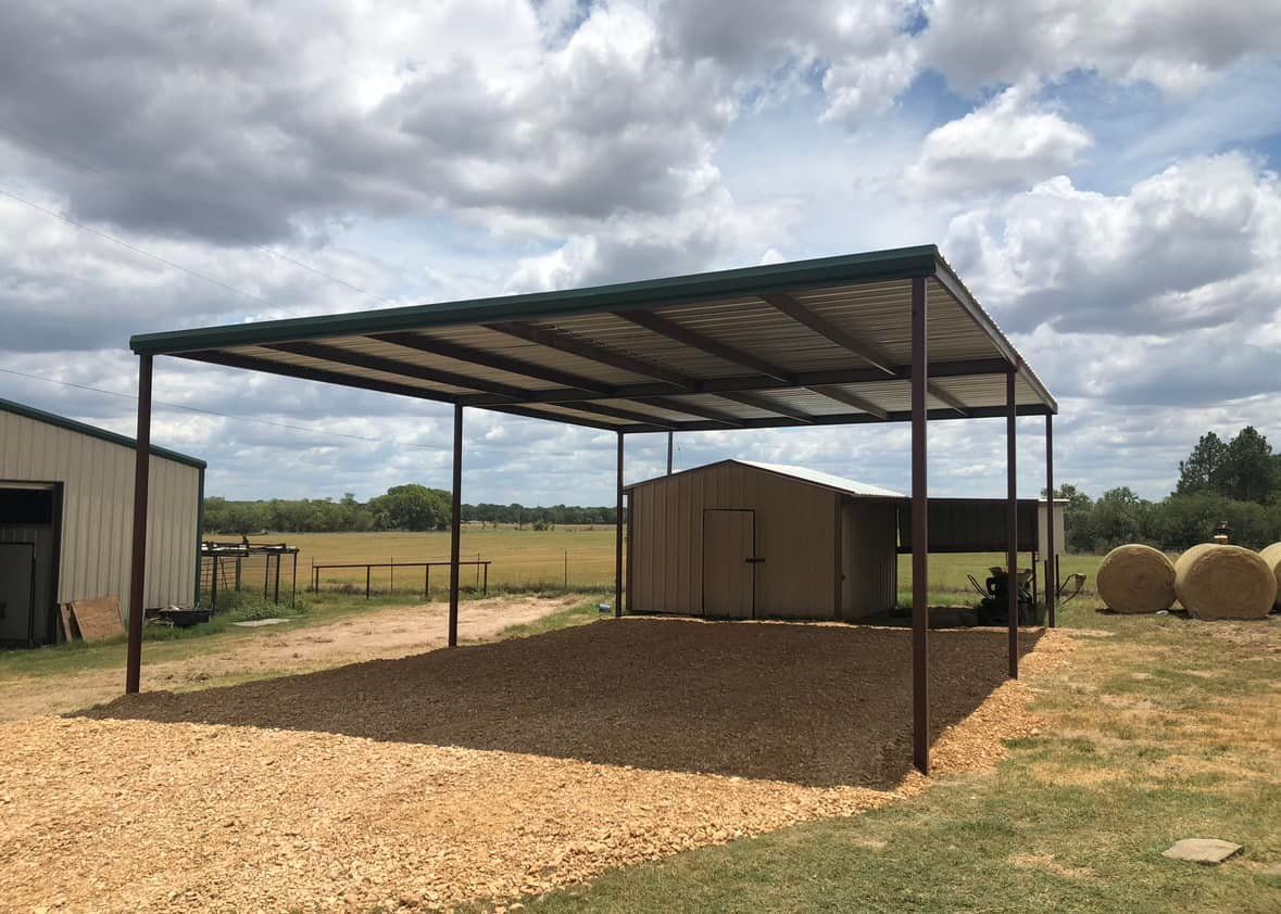 Open-air shelter with gravel base, metal roof, and brown metal supports, on a grassy field; cloudy sky.