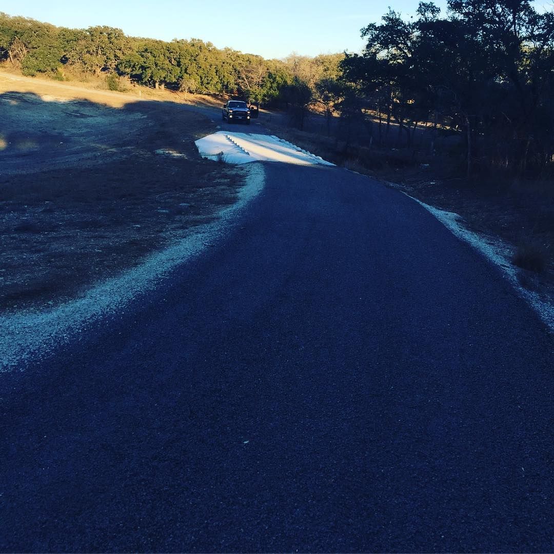 Dark asphalt road winding through a grassy landscape, car in the distance, blue sky.