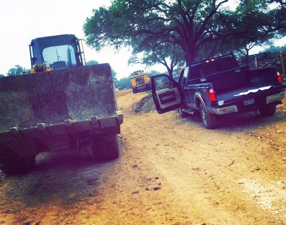 A black pickup truck parked next to a large front-end loader on a dirt road, with another loader in the distance.