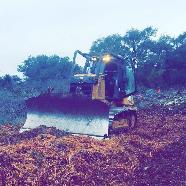 Bulldozer clearing debris on a dirt path, trees in the background, overcast sky.