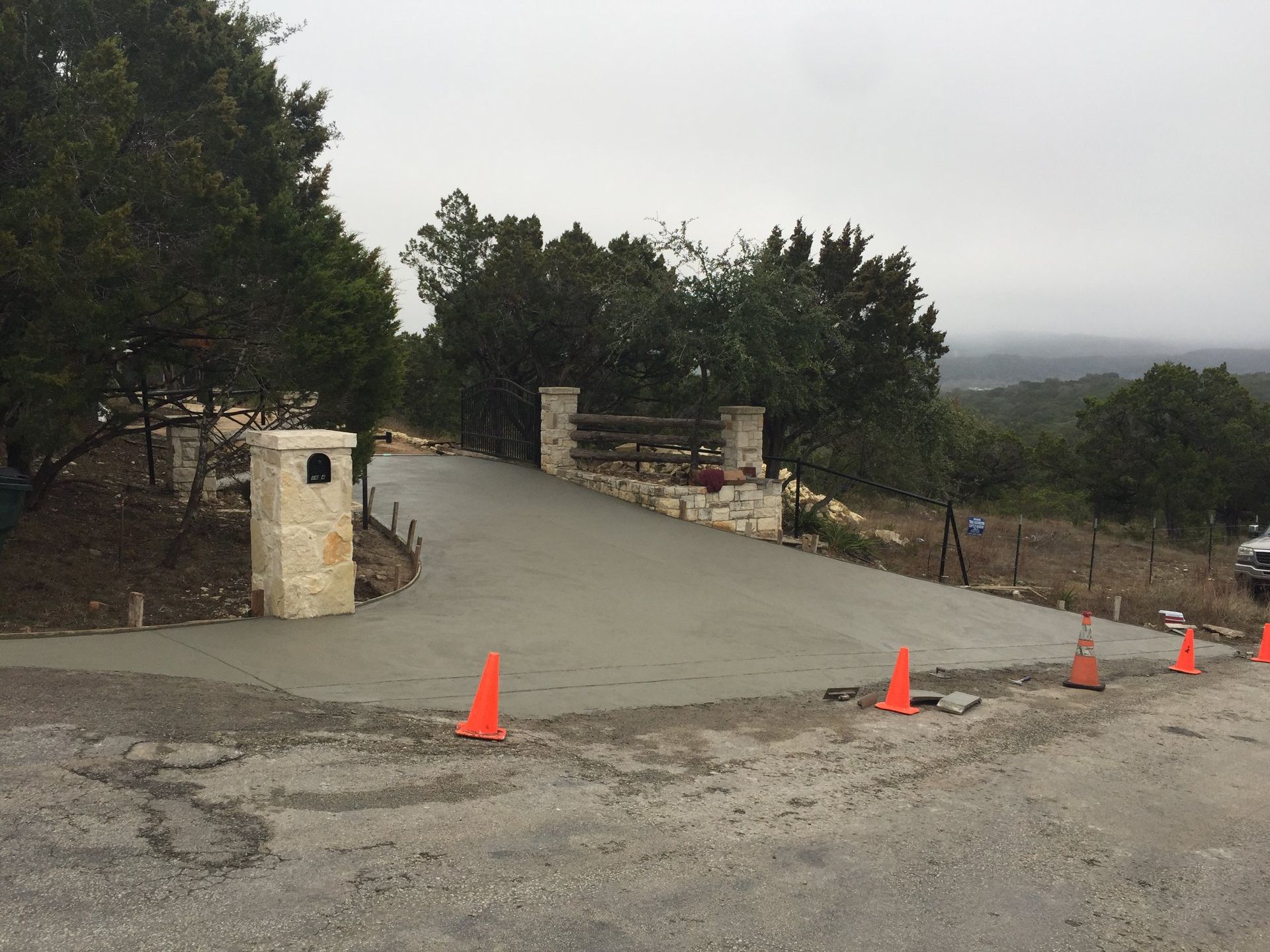 Freshly poured concrete driveway leading to a stone entrance with trees in the background. Orange cones mark the edges.