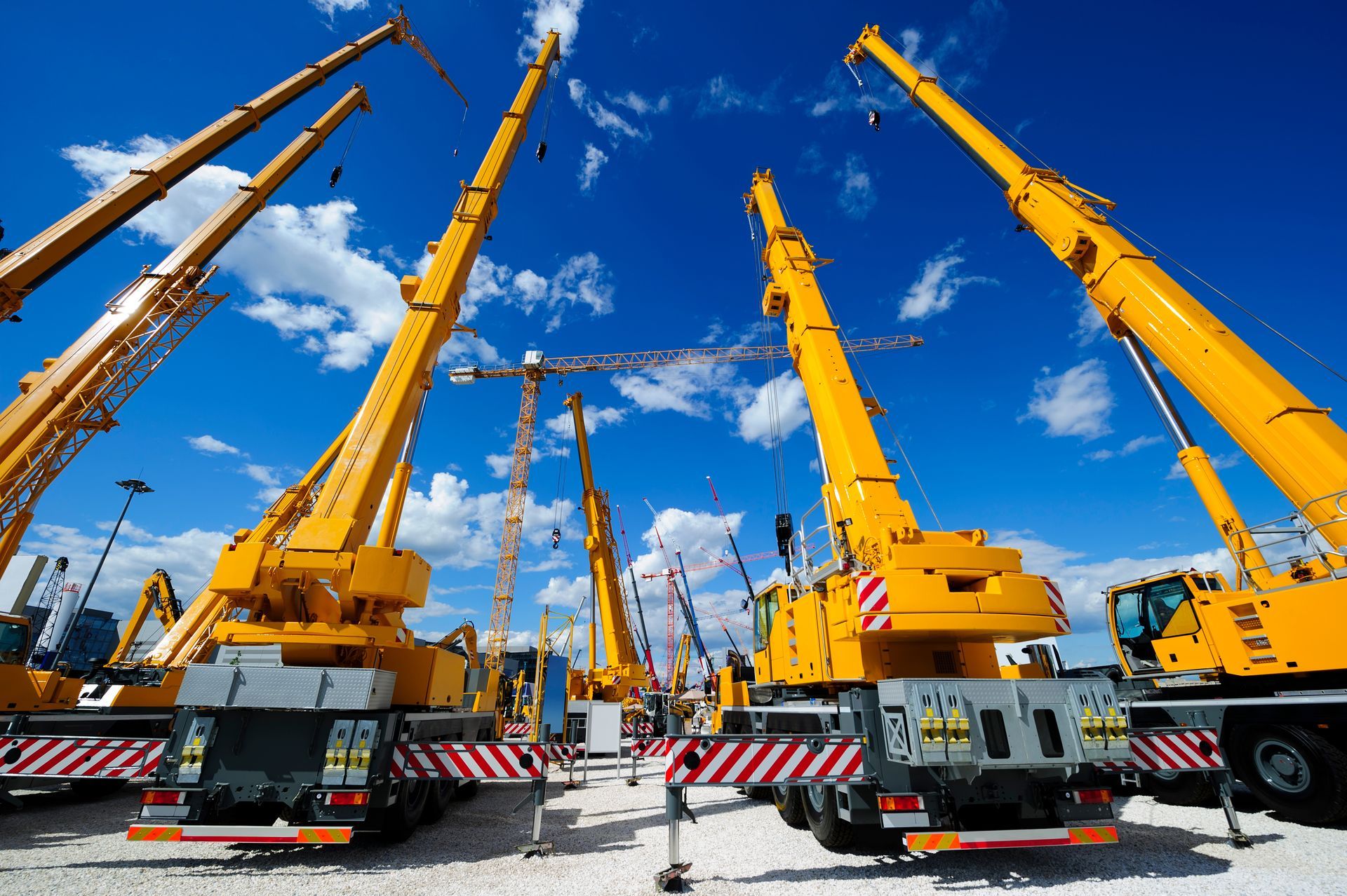 Multiple large construction cranes positioned on a gravel surface under a blue sky with scattered clouds.