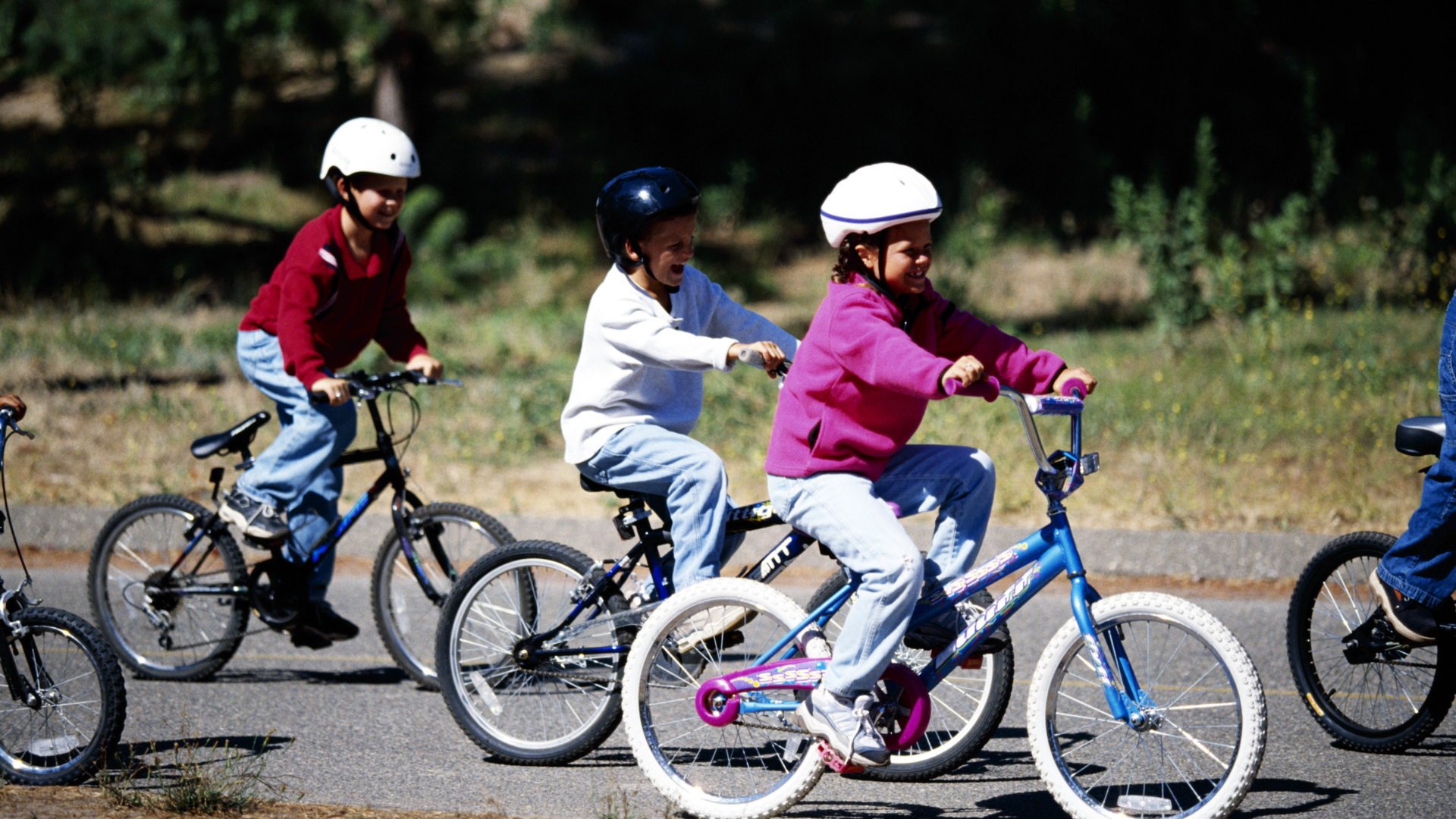 Three children on bikes, wearing helmets, riding on a paved path outdoors.