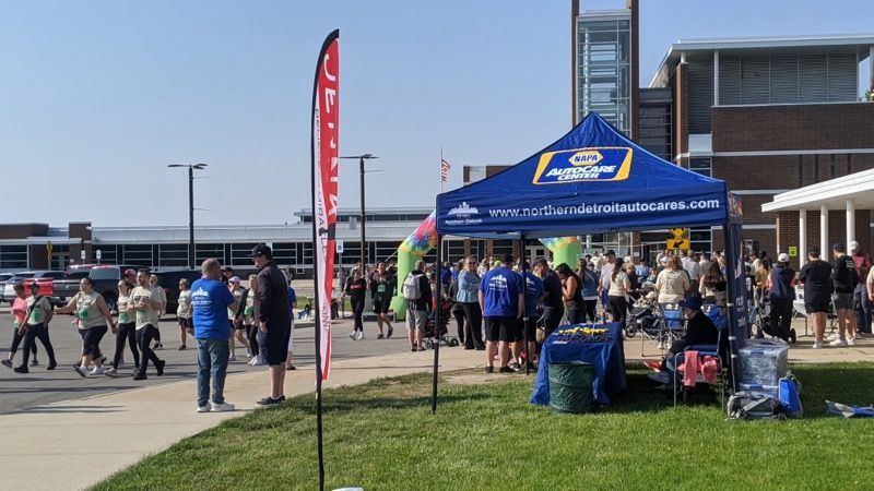 Crowd gathers near an NAPA AutoCare tent at an outdoor event near a large building. People walk and stand in sunlight.