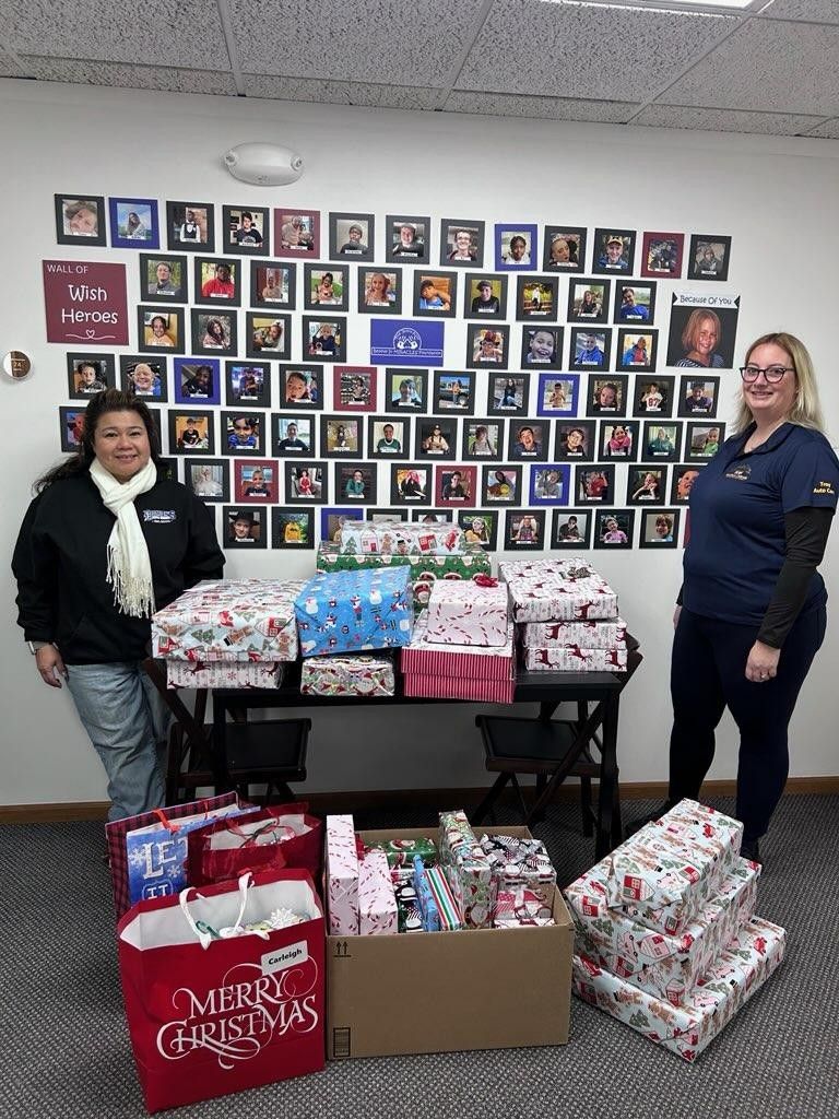 Two women stand with many wrapped gifts. A wall with framed photos is behind them.