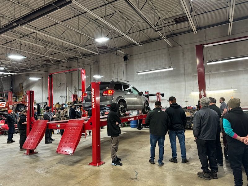 Group observing car on a lift in a garage. Red lift, concrete floor, overhead lights.