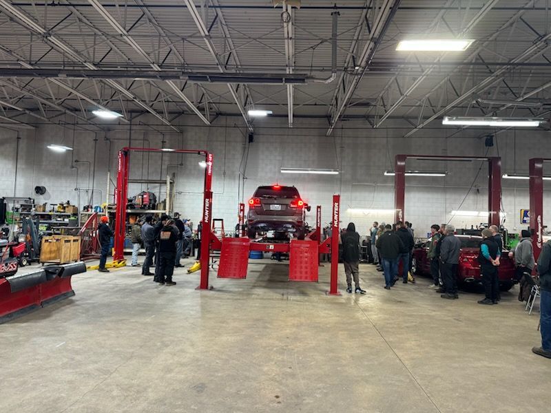Auto repair shop interior with vehicle on a lift, people observing, red equipment and concrete floor.
