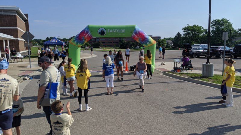 People walking/running toward finish line arch at an outdoor event. Green arch with tie-dye, people, blue sky.