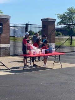 People at a red table selling treats in front of a brick fence, on a sunny day.