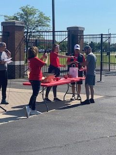 People at a table outside, handing out items. Red table and shirts, black fence, brick structure.