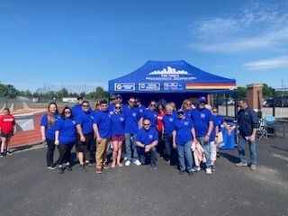 Group of people in blue shirts pose in front of a blue tent, outdoors on a sunny day.