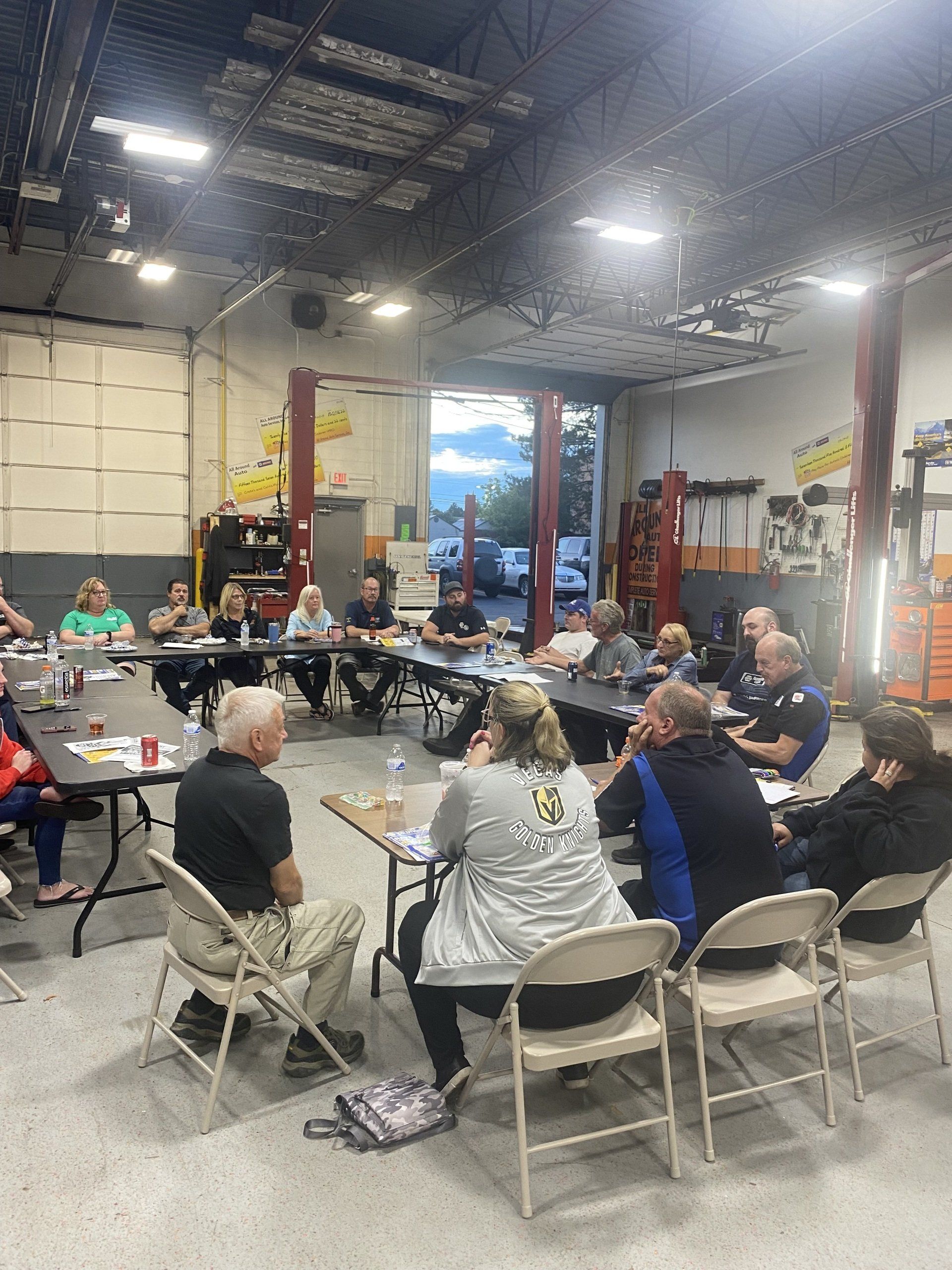 Group seated around tables in a garage, appearing to hold a meeting.