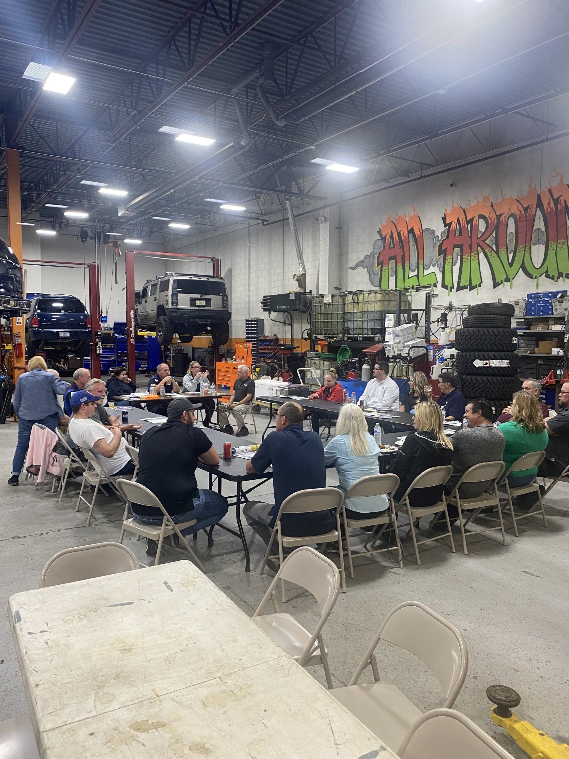 Group of people seated at tables, likely in a garage, having a meeting.