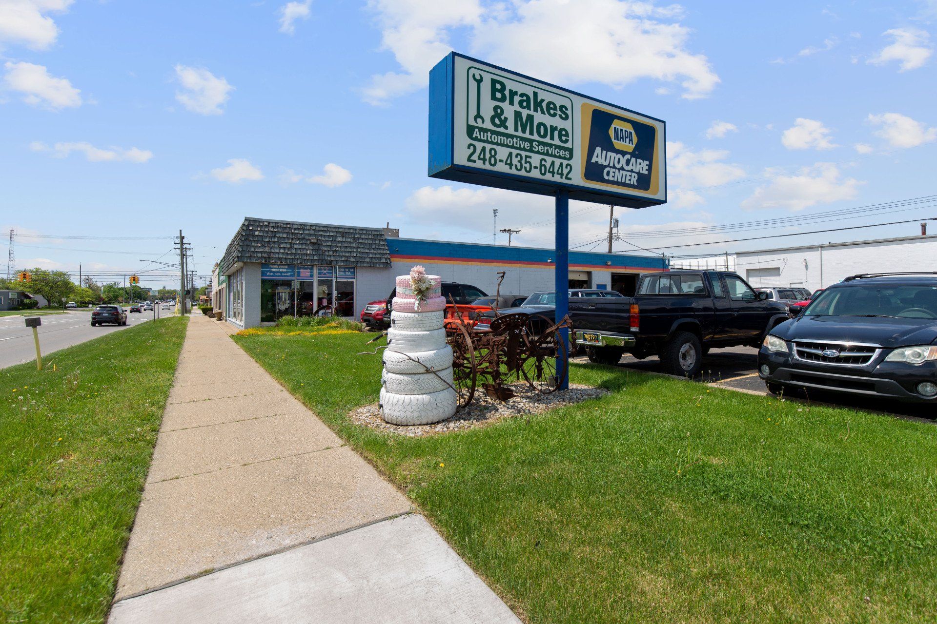 Brakes & More auto repair shop sign, brick building, cars parked in front, grass and sidewalk along a road.