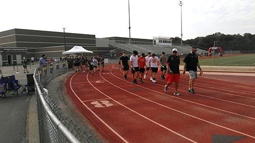 Runners on a red track at a stadium, preparing to start or cooling down. A tent and building are in the background.