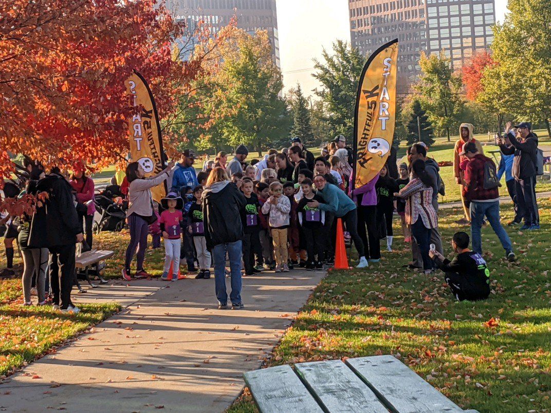 People gather for an outdoor event with orange and yellow banners, trees, and a pathway.