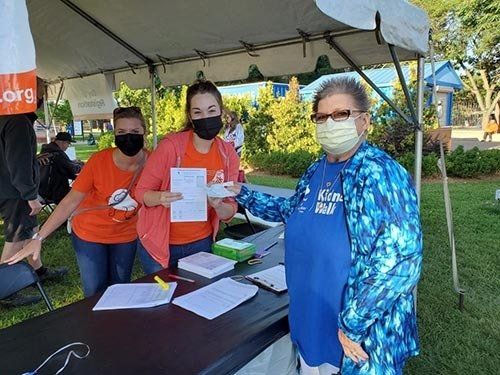 Three masked people at a table; one hands document to a person in a blue shirt under a tent, outdoors.