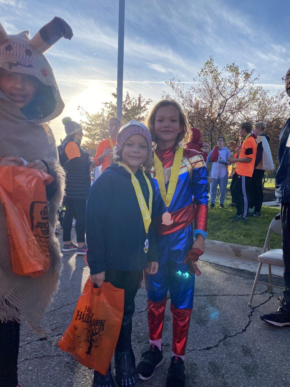 Two children in costumes pose with medals and orange bags outdoors at sunset.